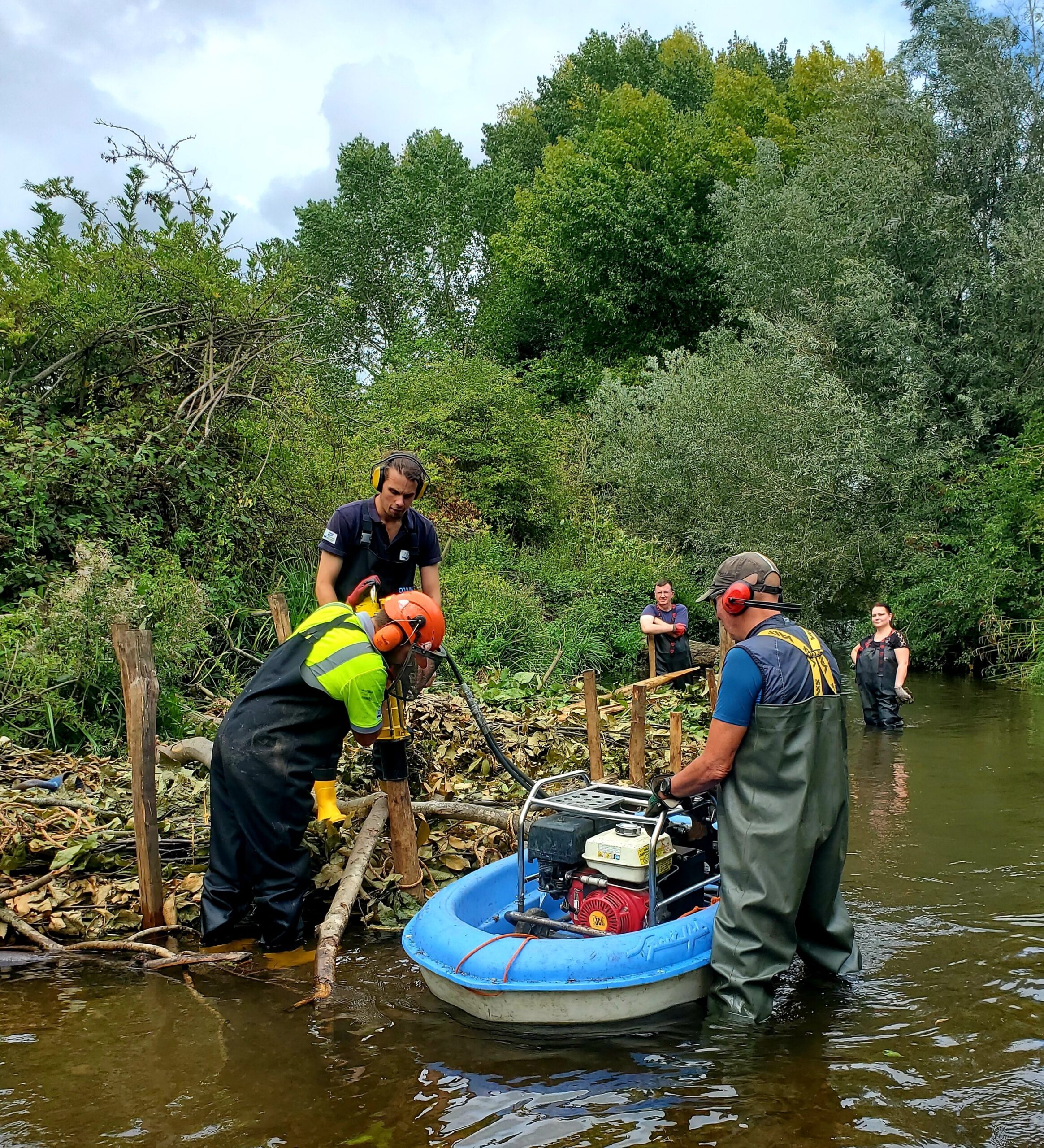 A group of people standing in a river alongside a shelf of branches and wooden posts.