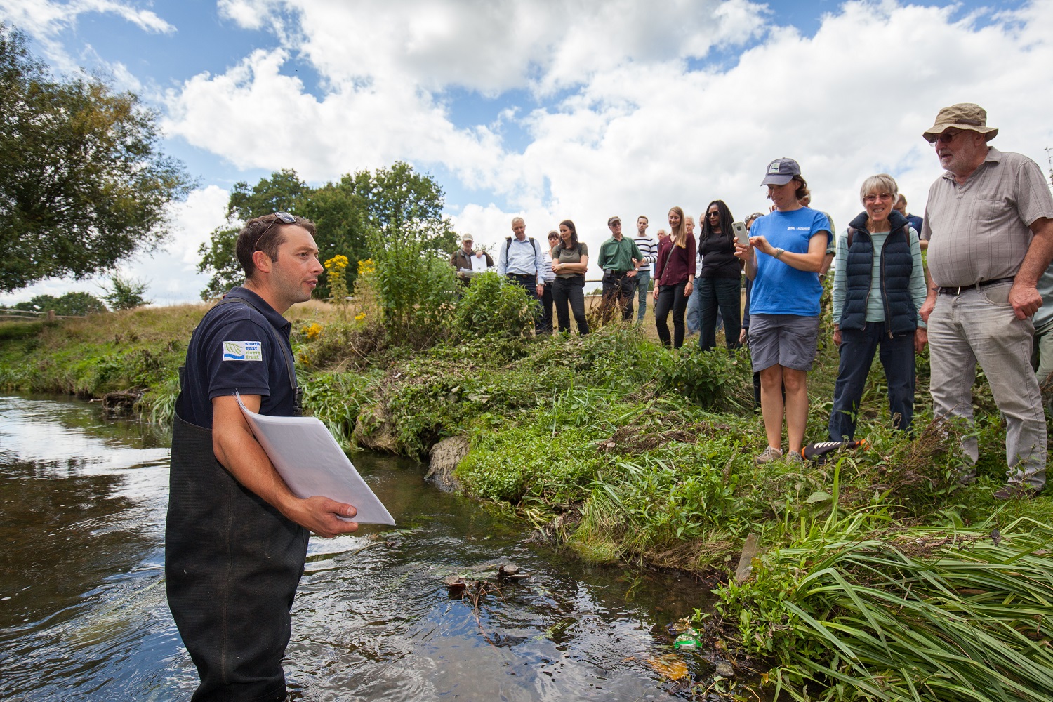 A man stands in a river, talking to people on the riverbank.