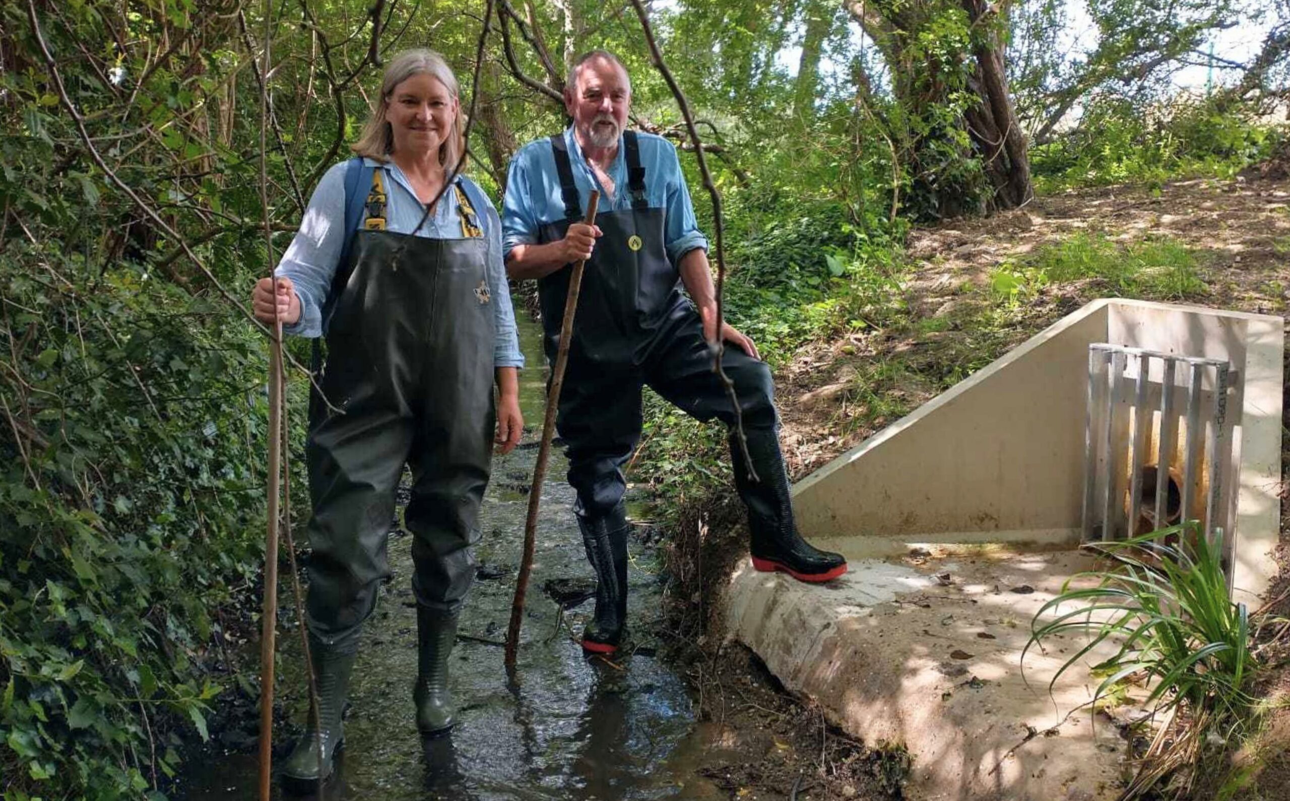 Two people with waders on standing in front of an outfall along a river bank.