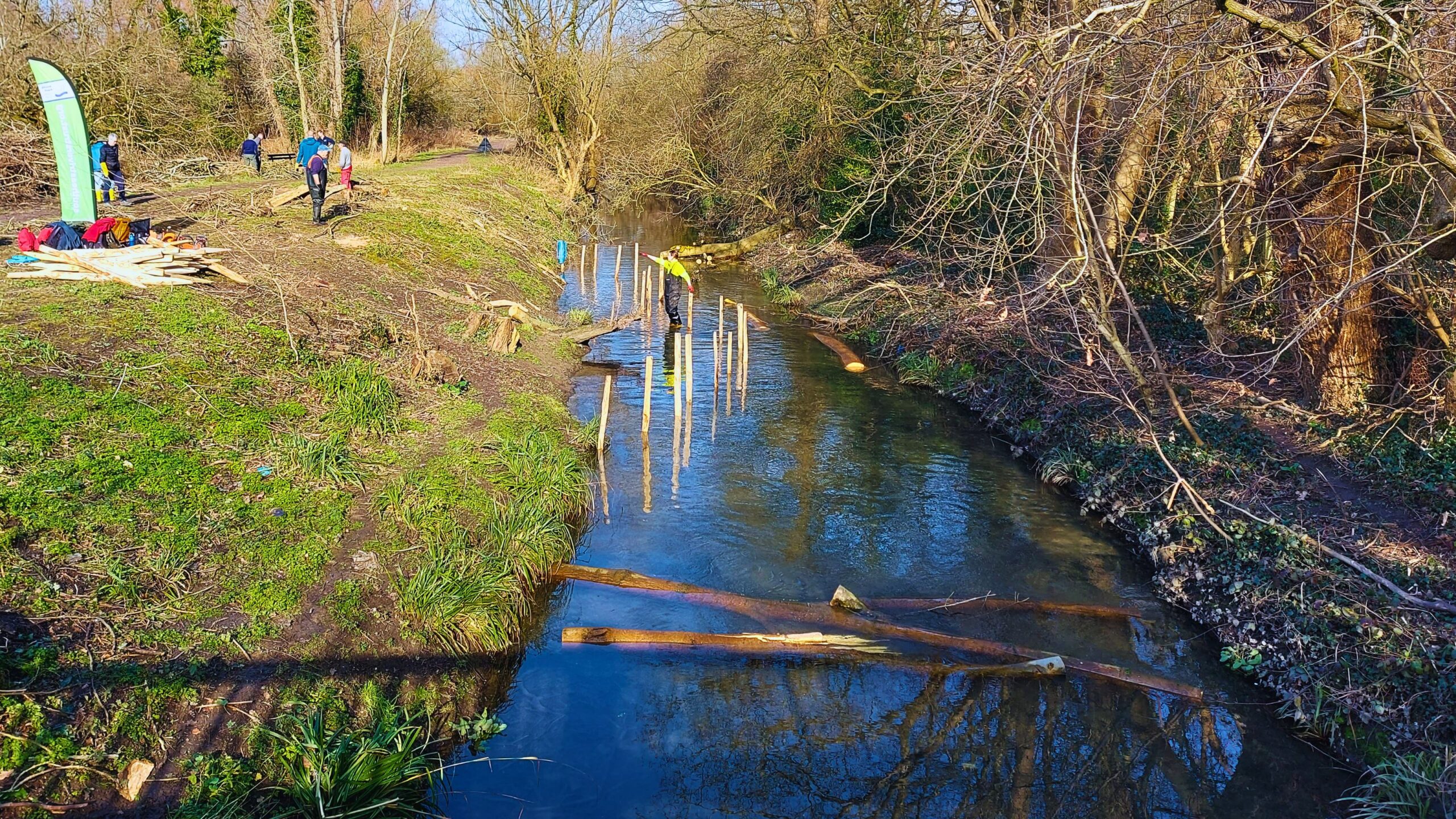 A view of a river with pinned tree trunks across the channel and wooden posts forming a semi-circle along the bank.