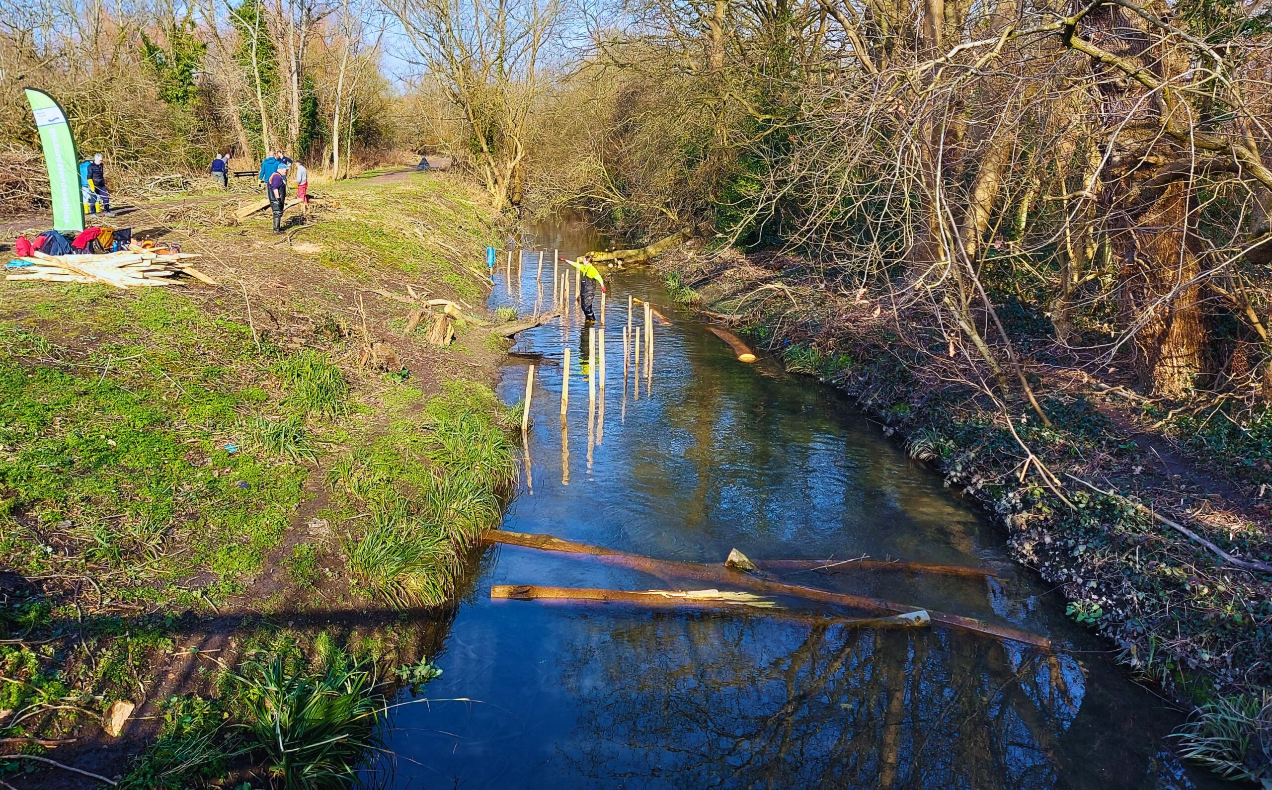 A view of a river with pinned tree trunks across the channel and wooden posts forming a semi-circle along the bank.