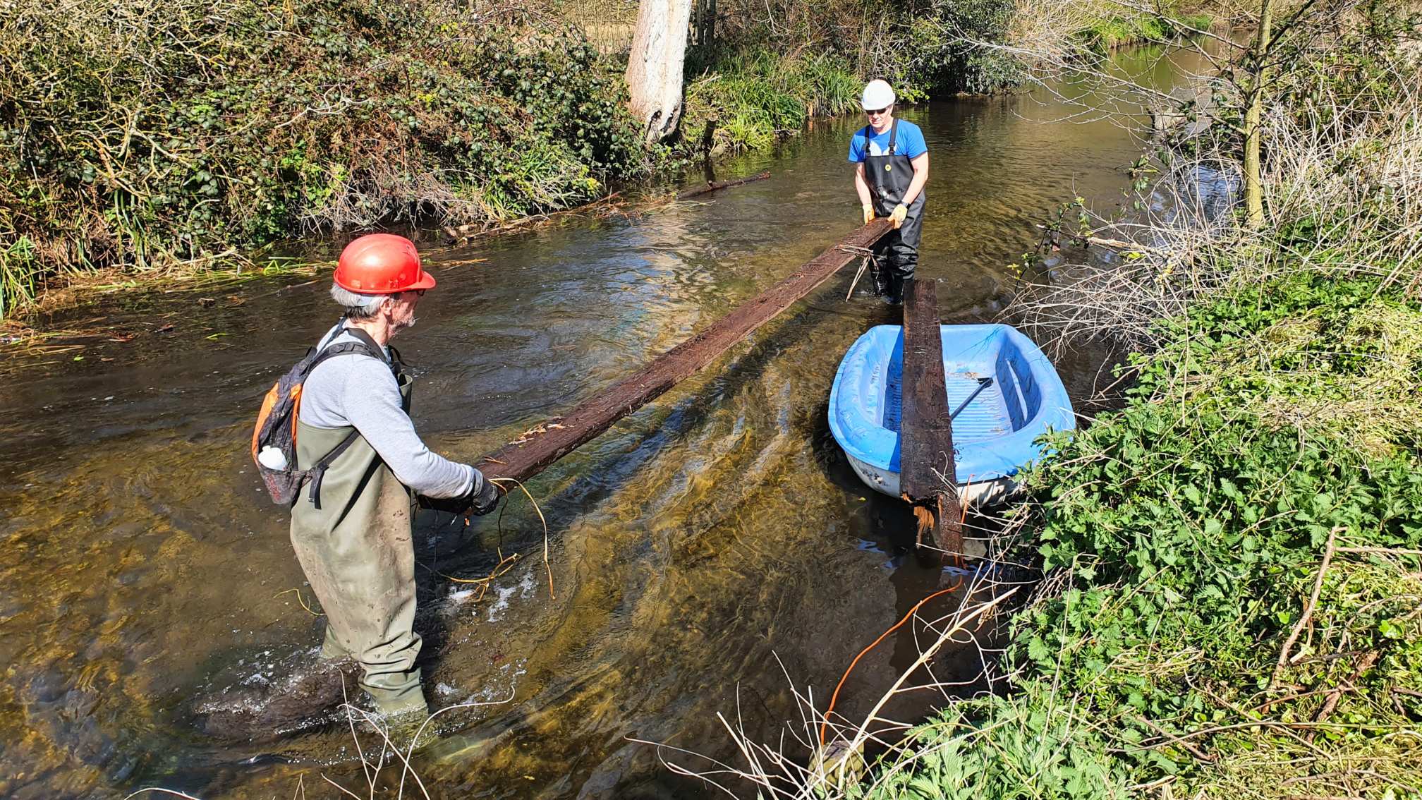 Two people standing in a river holding a large wooden plank with a small blue boat alongside them.