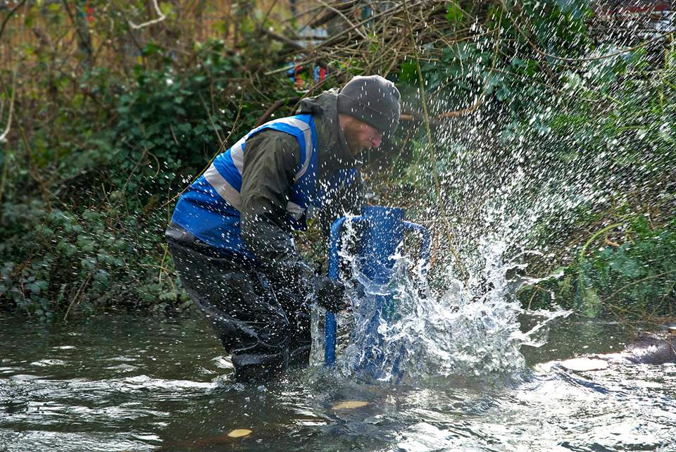 A man in a blue high vis vest standing in water using a post driver.