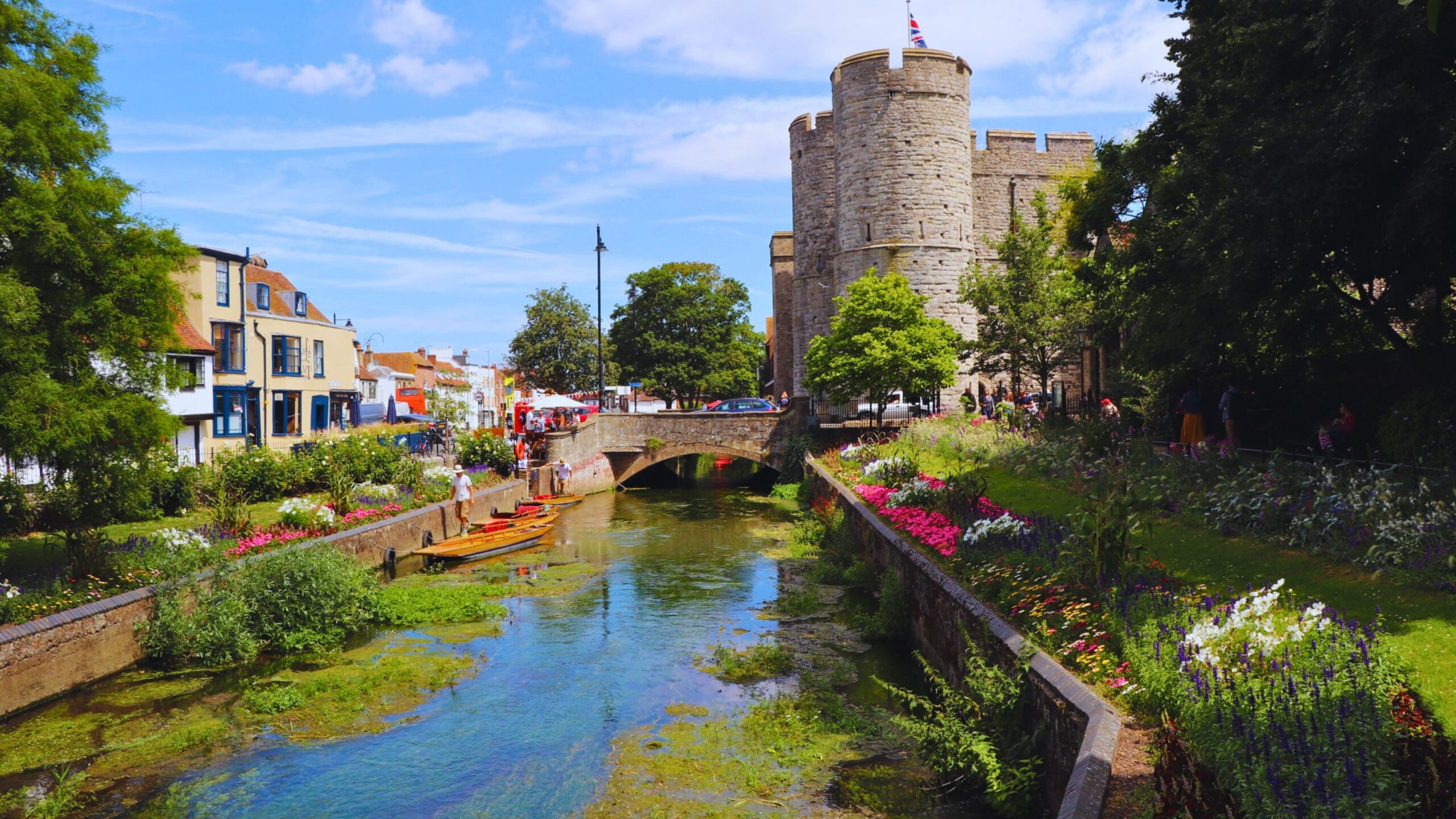 A view of a river flowing through a town with a row of shops one side and a castle the other side.