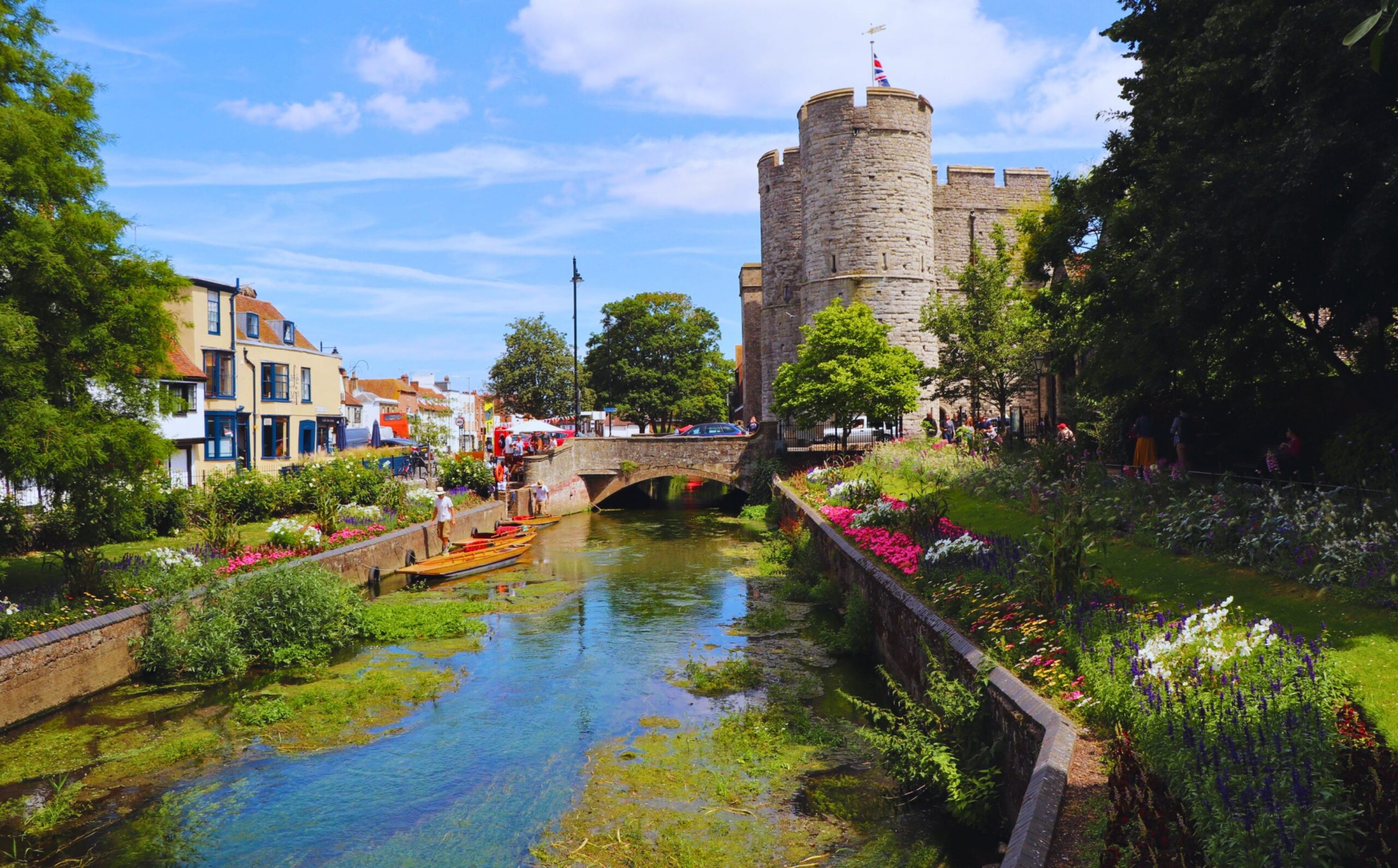 A view of a river flowing through a town with a row of shops one side and a castle the other side.