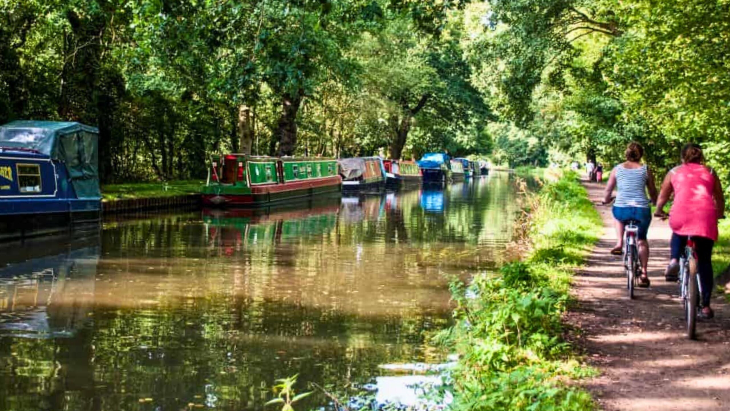 The River Wey tow path with cyclists on the bank and canal boats on the other side.