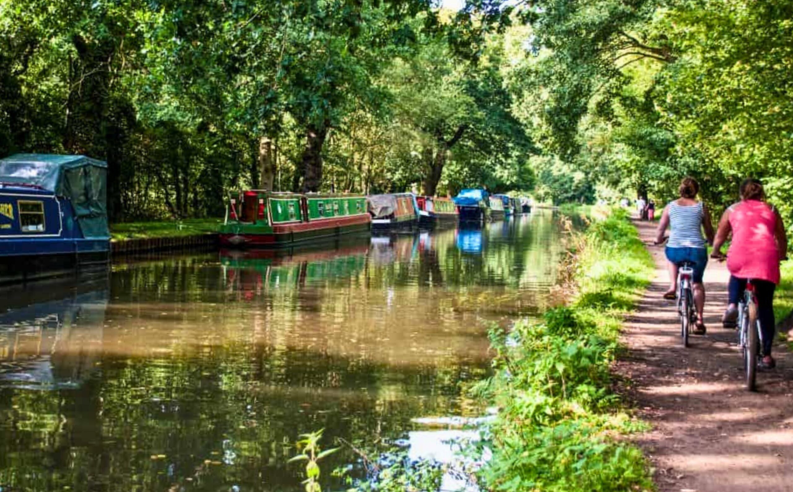 The River Wey tow path with cyclists on the bank and canal boats on the other side.