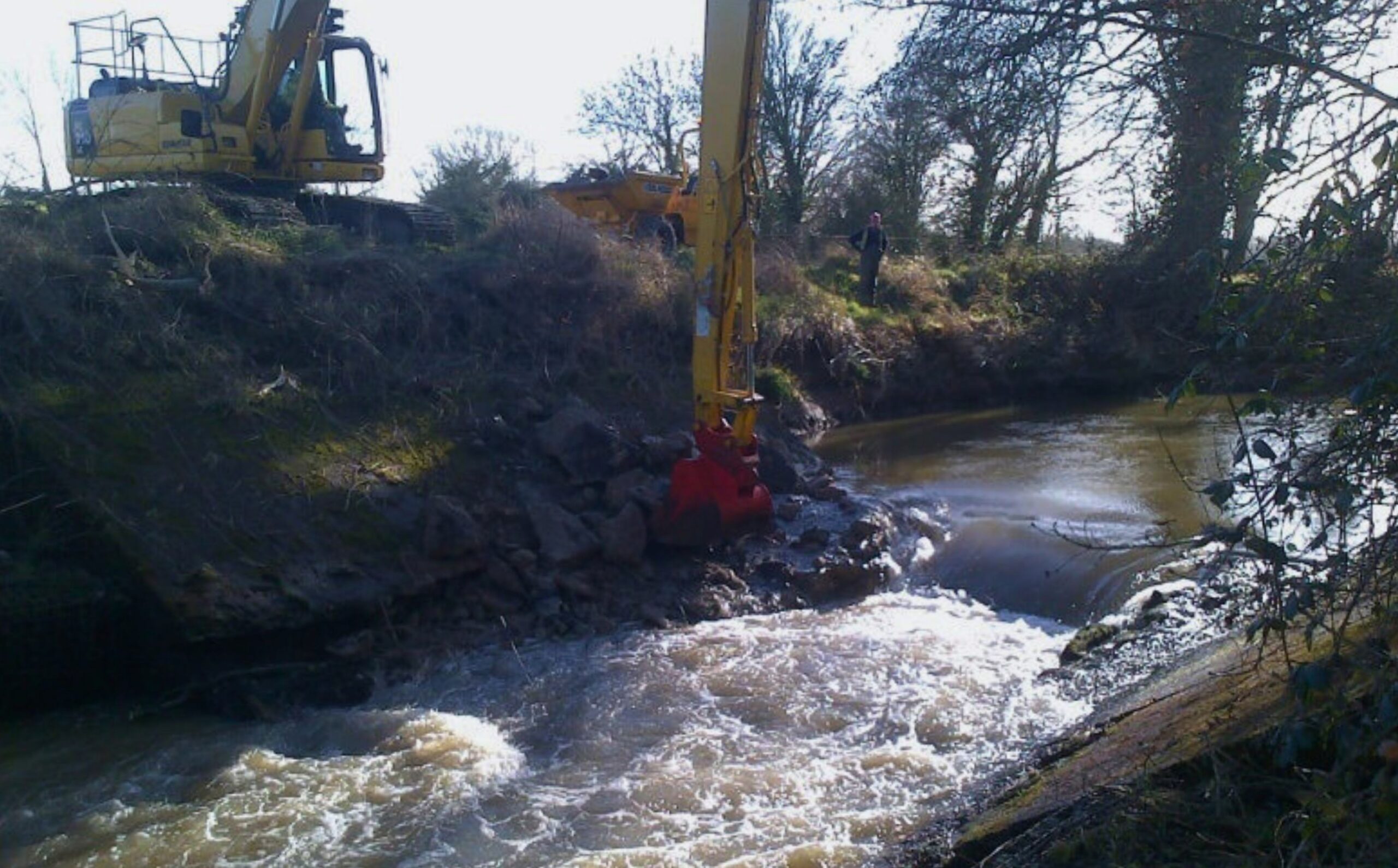 A digger on a river bank drilling into a concrete weir.