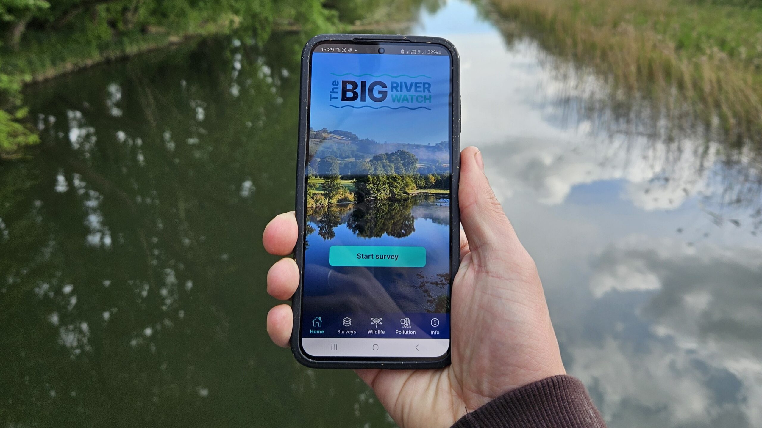 A hand holding a mobile phone in front of a river with the Big River Watch app on the screen.