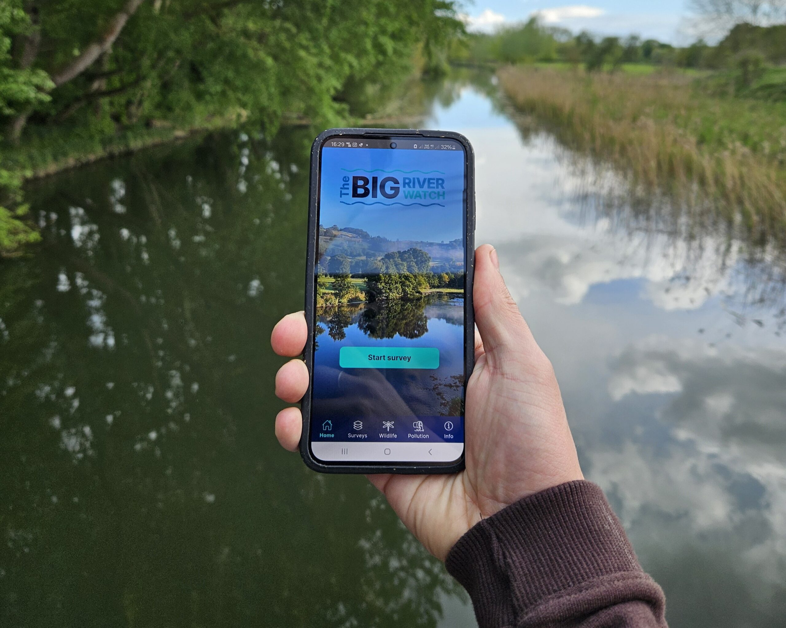 A hand holding a mobile phone in front of a river with the Big River Watch app on the screen.