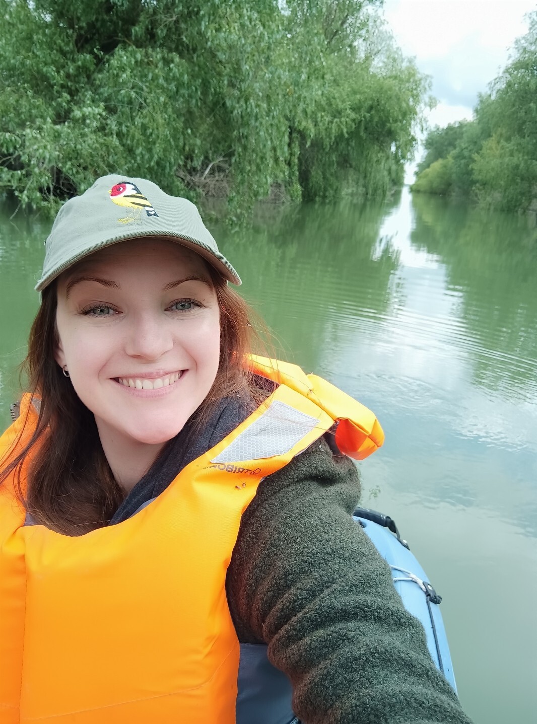 A woman wearing an orange high vis vest and green baseball cap, whilst sitting in a boat on a river.