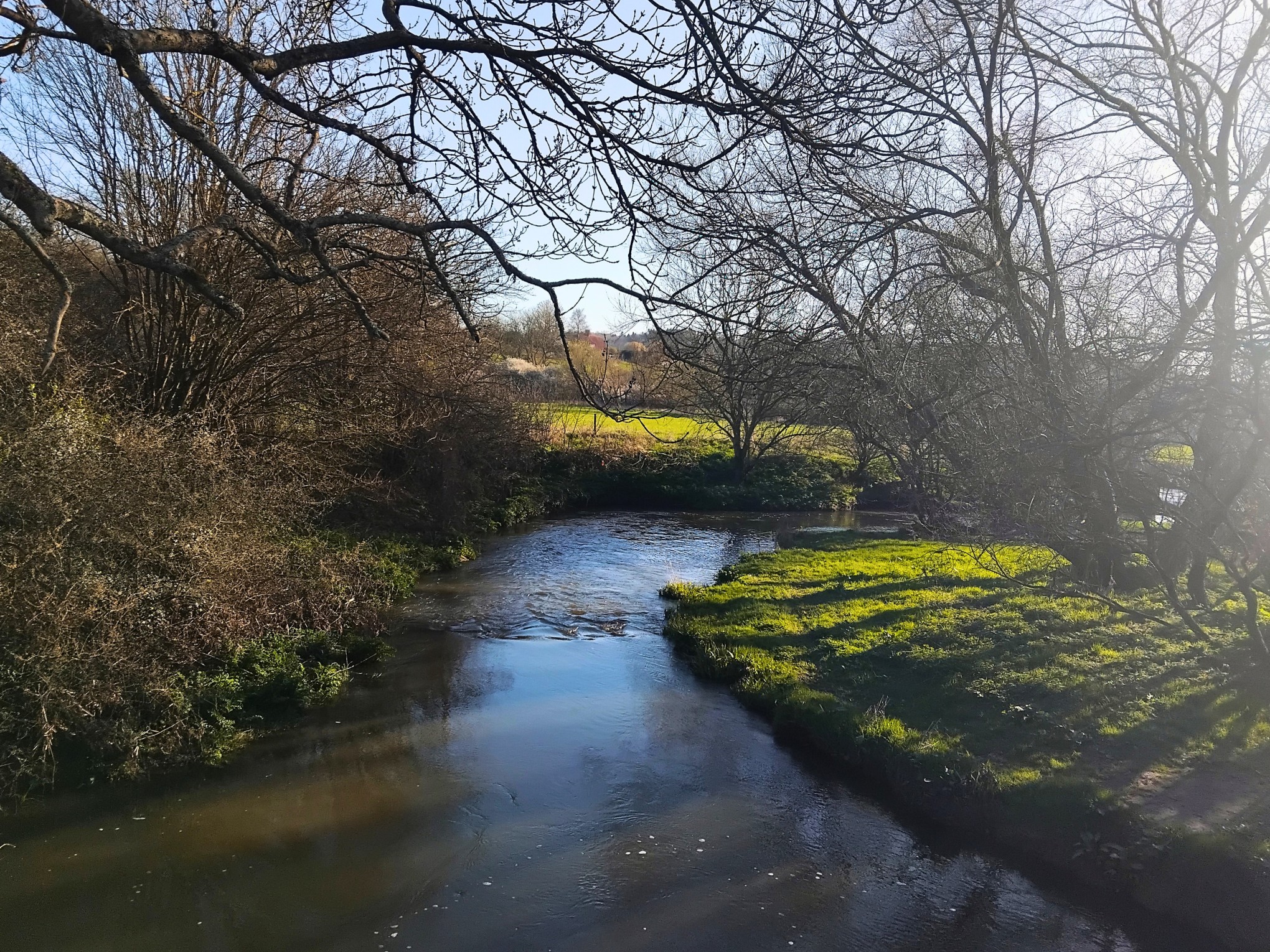 A river running through farmland with fields in the distance and trees either side of the bank.