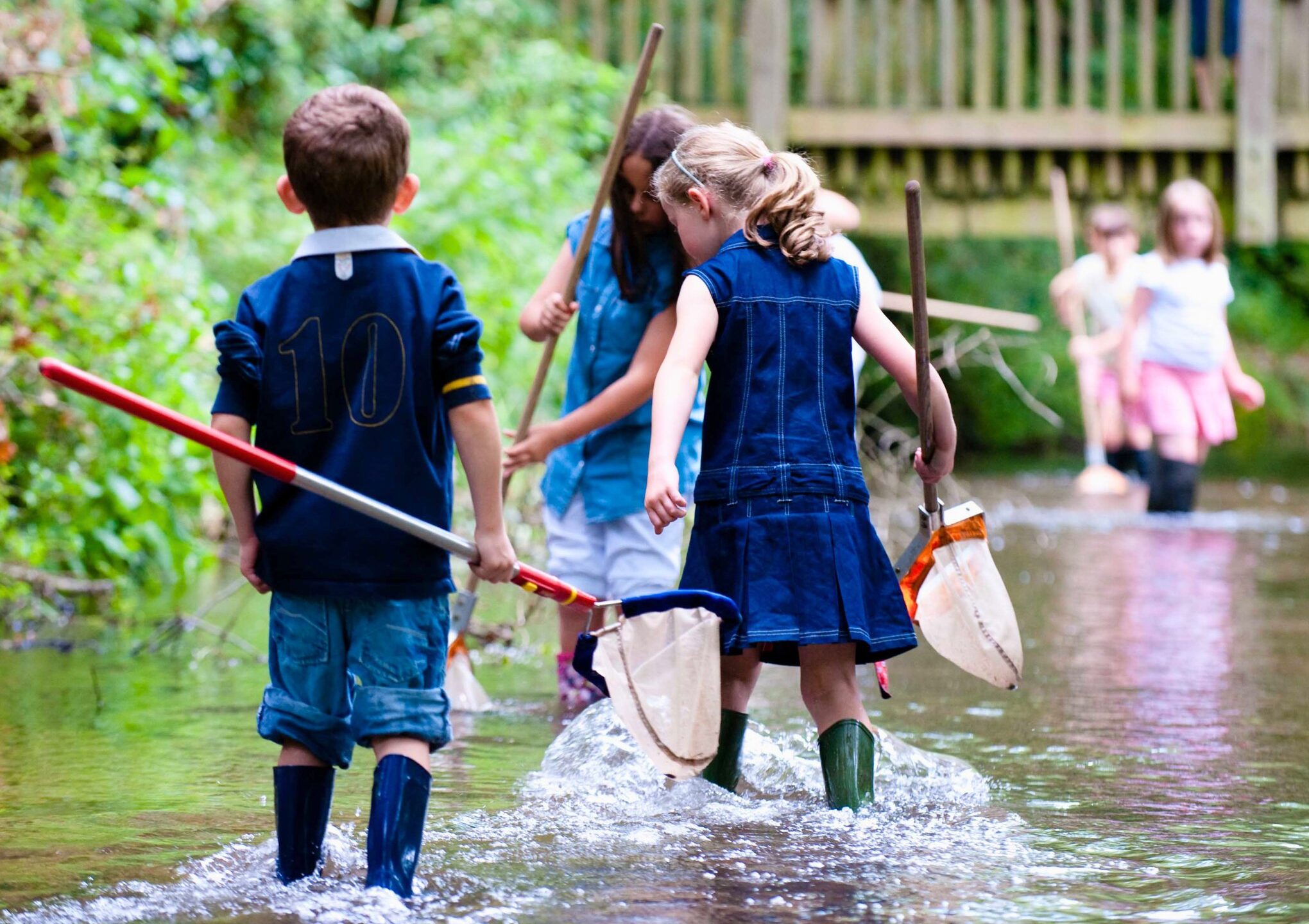Children holding nets and wearing wellies walking through a river.