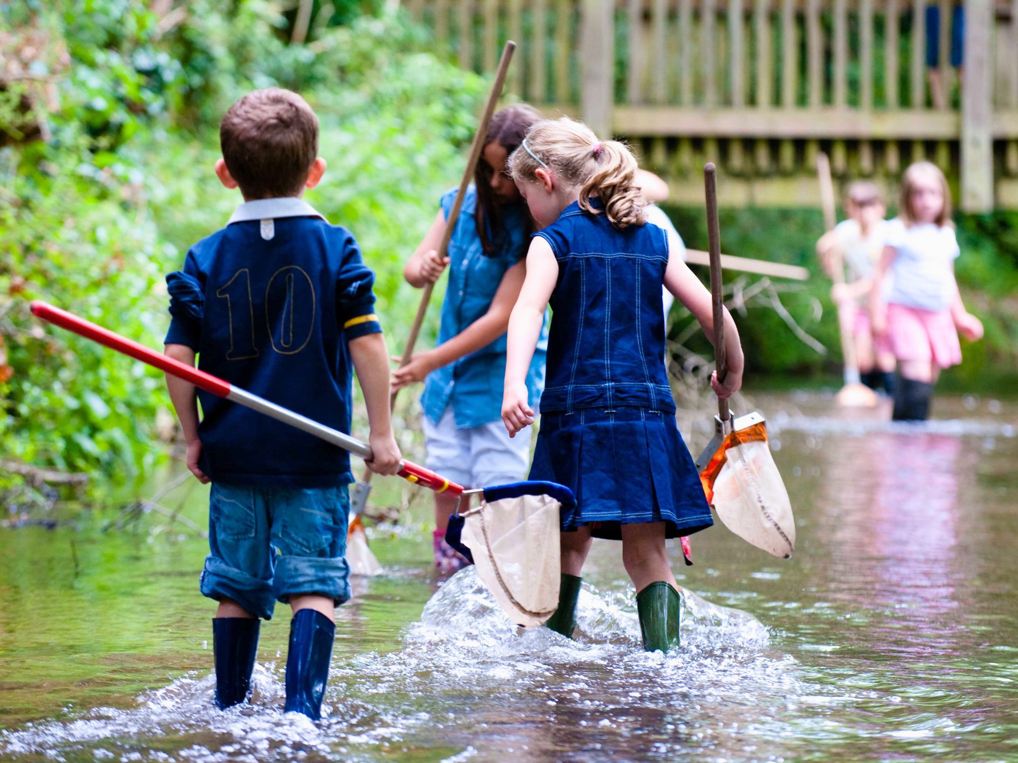 Children holding nets and wearing wellies walking through a river.