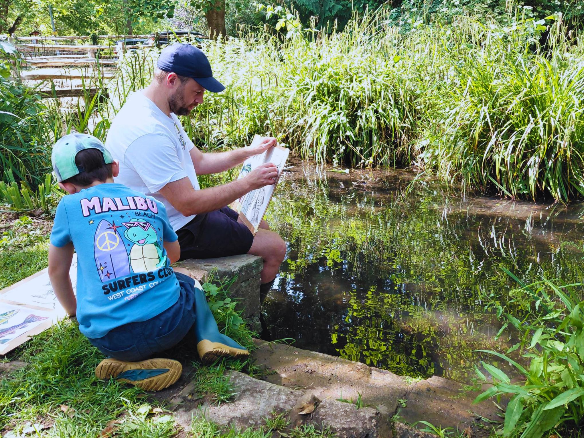 A man and a young boy sit along side a lake whilst painting a picture.