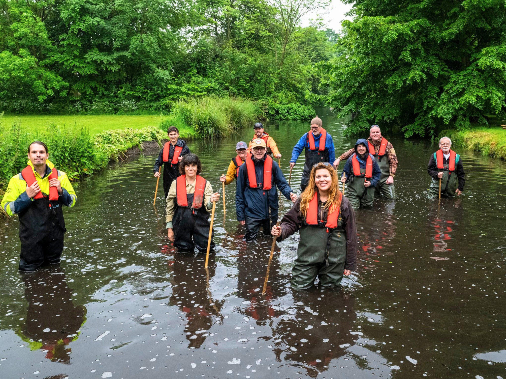 A group of people wearing life jackets and waders whilst standing in the middle of a river holding wooden poles.