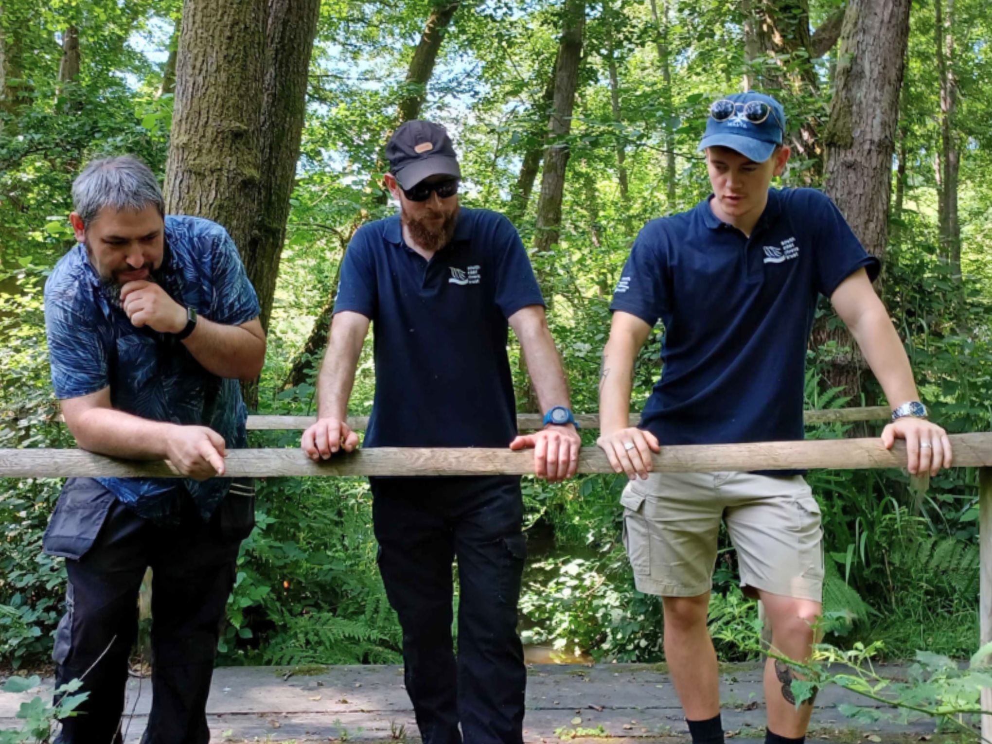 Three men standing on a wooden bridge over a stream in a woodland.