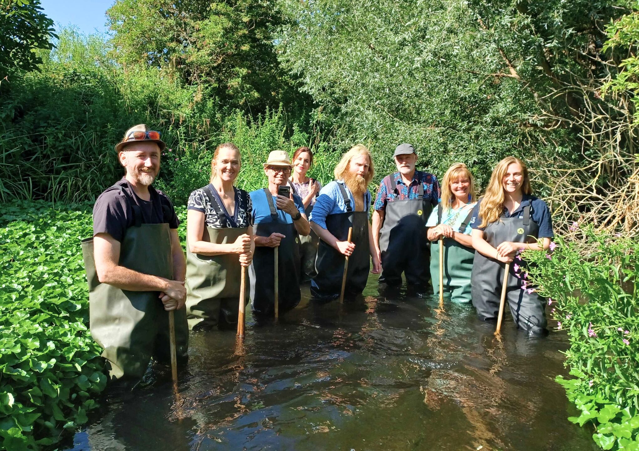 A group of people standing in a river wearing waders and holding wooden poles.