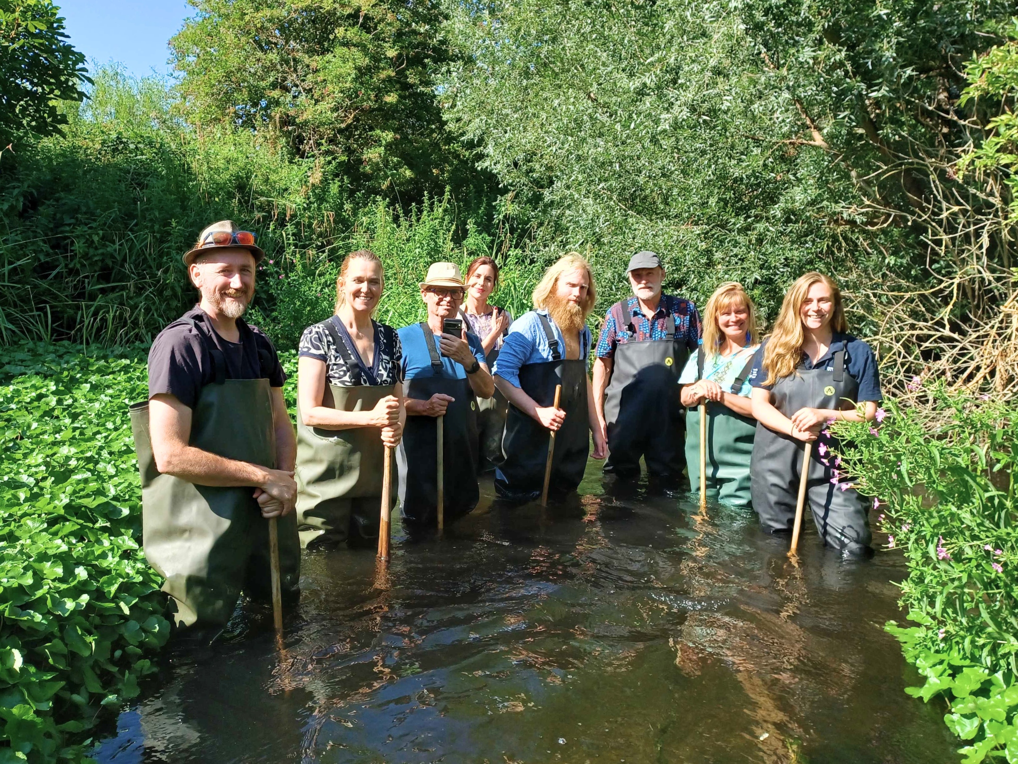A group of people standing in a river wearing waders and holding wooden poles.