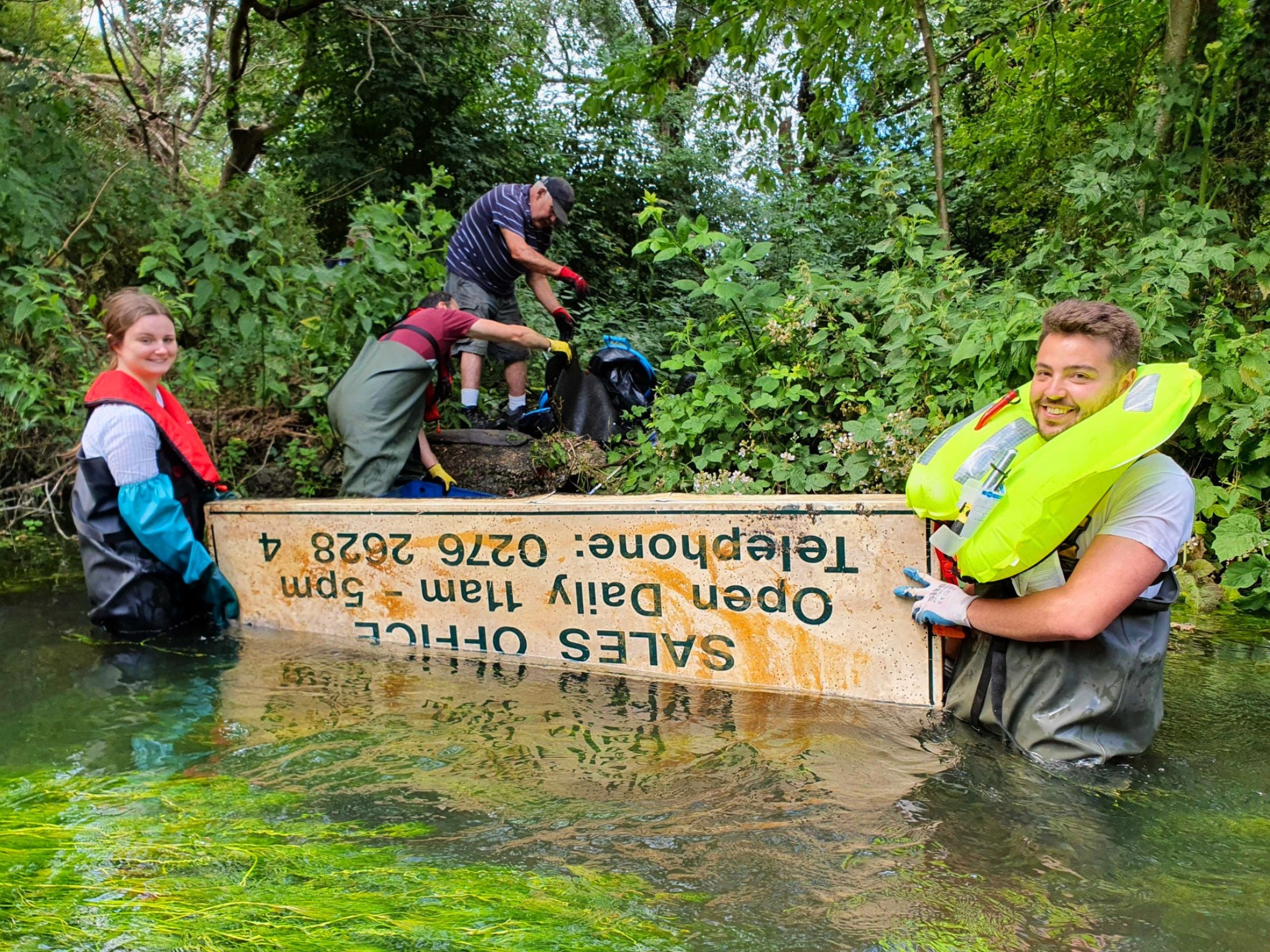 Two volunteers in waders and life jackets standing in a river holding a sign found in the river during a litter pick event. Two more people on the riverbank in the background sorting out waders.