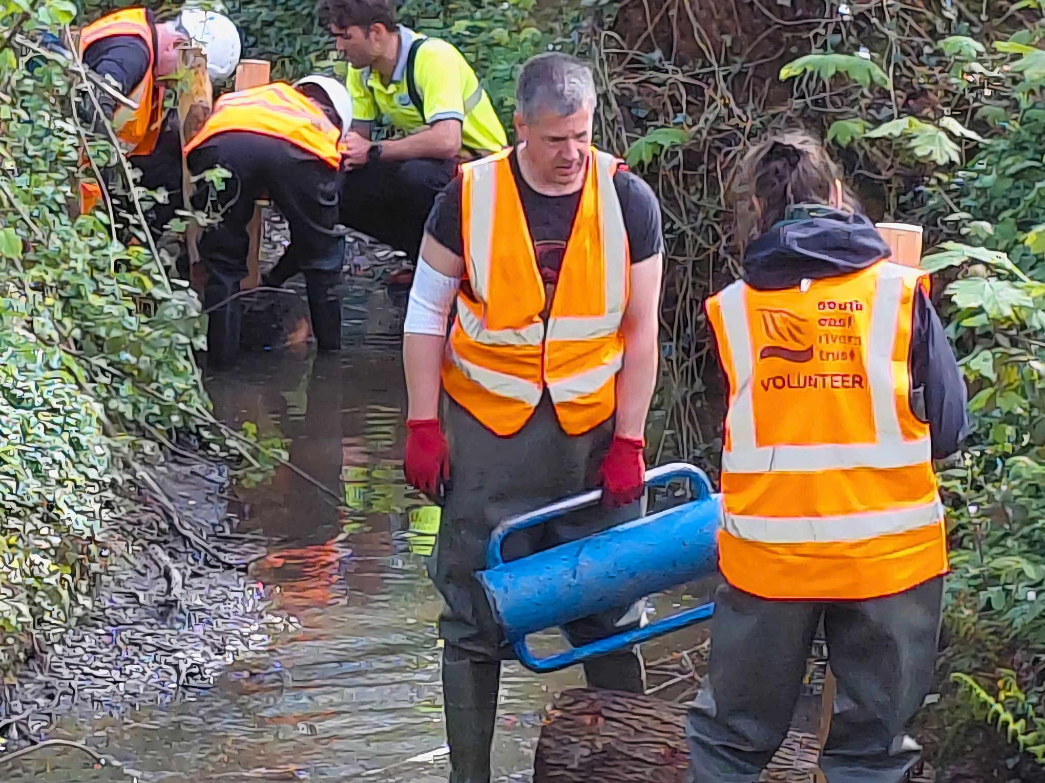 People in high vis vests working in a river channel, using post hammers to drive wooden posts into the ground.