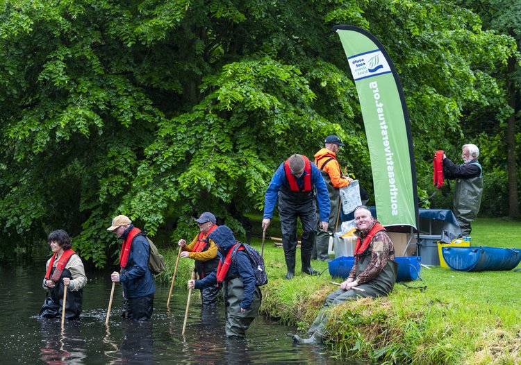 People in high vis vests climbing into a river and standing along the river bank. A South East Rivers Trust pop up banner is on the river bank.