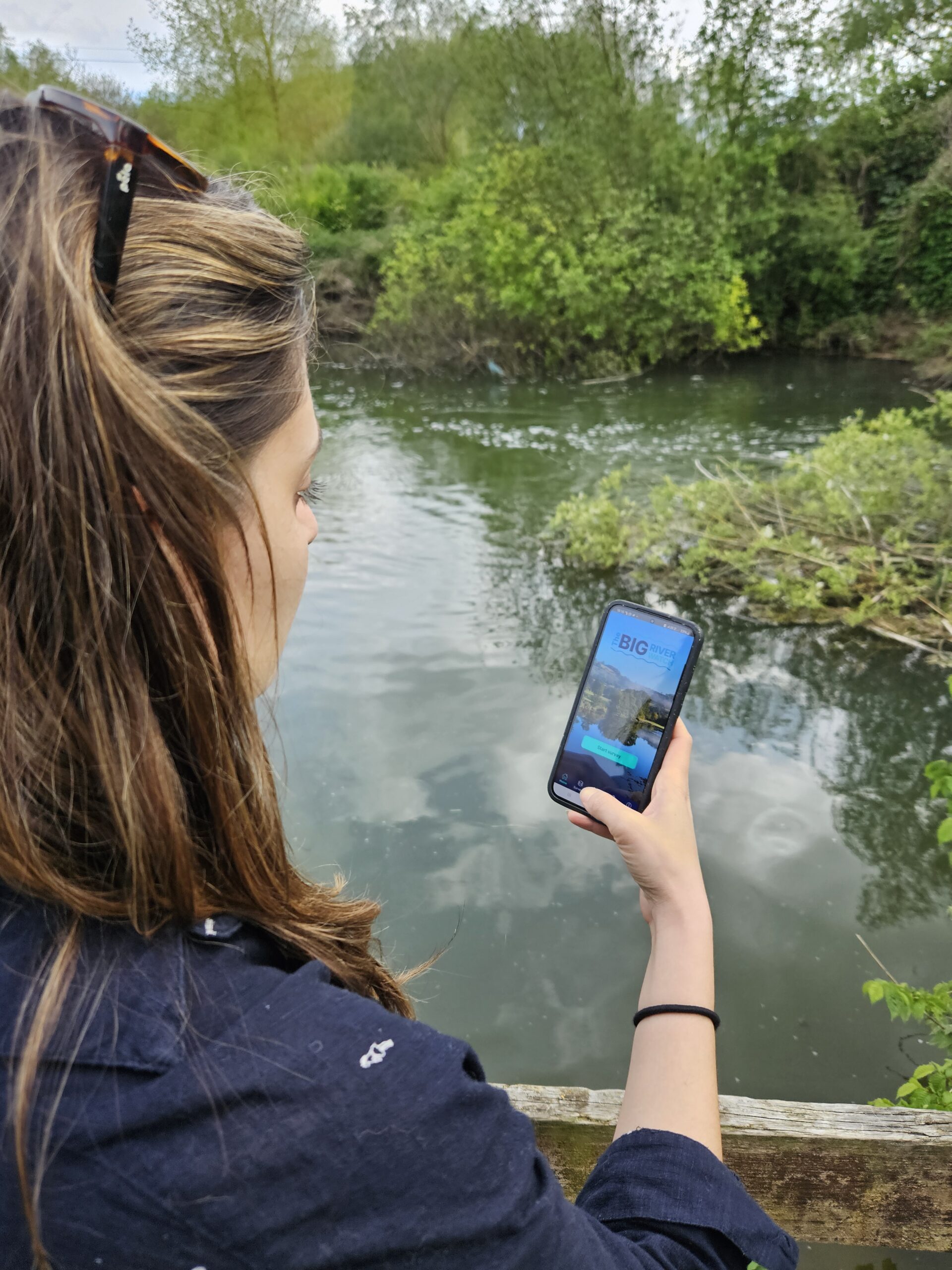 A person holding a mobile phone in front of a river.